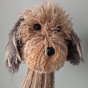 Puede incluir: Una funda para palo de golf con forma de perro, hecha a mano con hilo texturizado en tonos beige y marrón. La funda presenta una cara peluda con ojos de plástico oscuro y una nariz negra, unida a un cuello de punto acanalado color beige.
