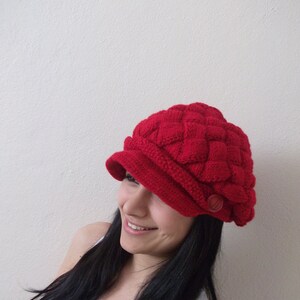May include: A woman with long black hair is wearing a red knitted hat with a button. The hat has a unique braided design.