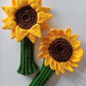Könnte beinhalten: Zwei gehäkelte Sonnenblumen-Golfschläger-Kopfbezüge. Die größere Sonnenblume ist gelb mit einer braunen Mitte und einem grünen Stiel. Die kleinere Sonnenblume ist ebenfalls gelb mit einer braunen Mitte und einem grünen Stiel.