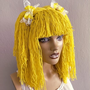 May include: A vibrant yellow wig with a fringe and two small bows. The wig is made of yarn and is styled on a mannequin head. The bows are yellow and white with lace trim. The mannequin is wearing a white top.