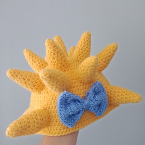 May include: A yellow crocheted hat with a spiky design and a baby blue bow. The hat is held in a hand.