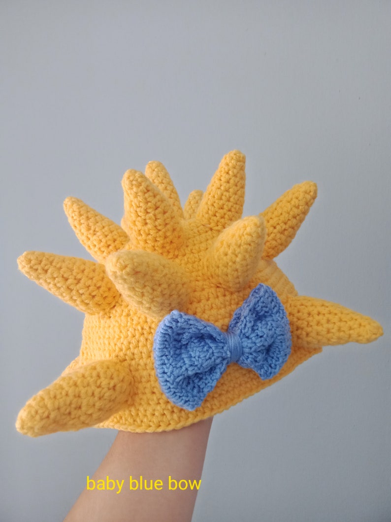 May include: A yellow crocheted hat with a spiky design and a baby blue bow. The hat is held in a hand.