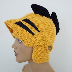 May include: A yellow crocheted hat shaped like a helmet with black stripes and a black crocheted crest. The hat has a flap that covers the ears and a button on the side.