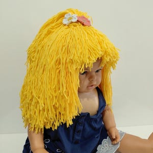May include: A doll wearing a handmade yellow yarn wig with a white and pink flower hair accessory. The doll is dressed in a blue dress with a white and blue striped trim.