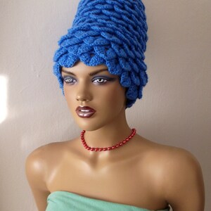 May include: A blue crocheted hat with a unique, pointed design. The hat is worn by a mannequin with a red beaded necklace.