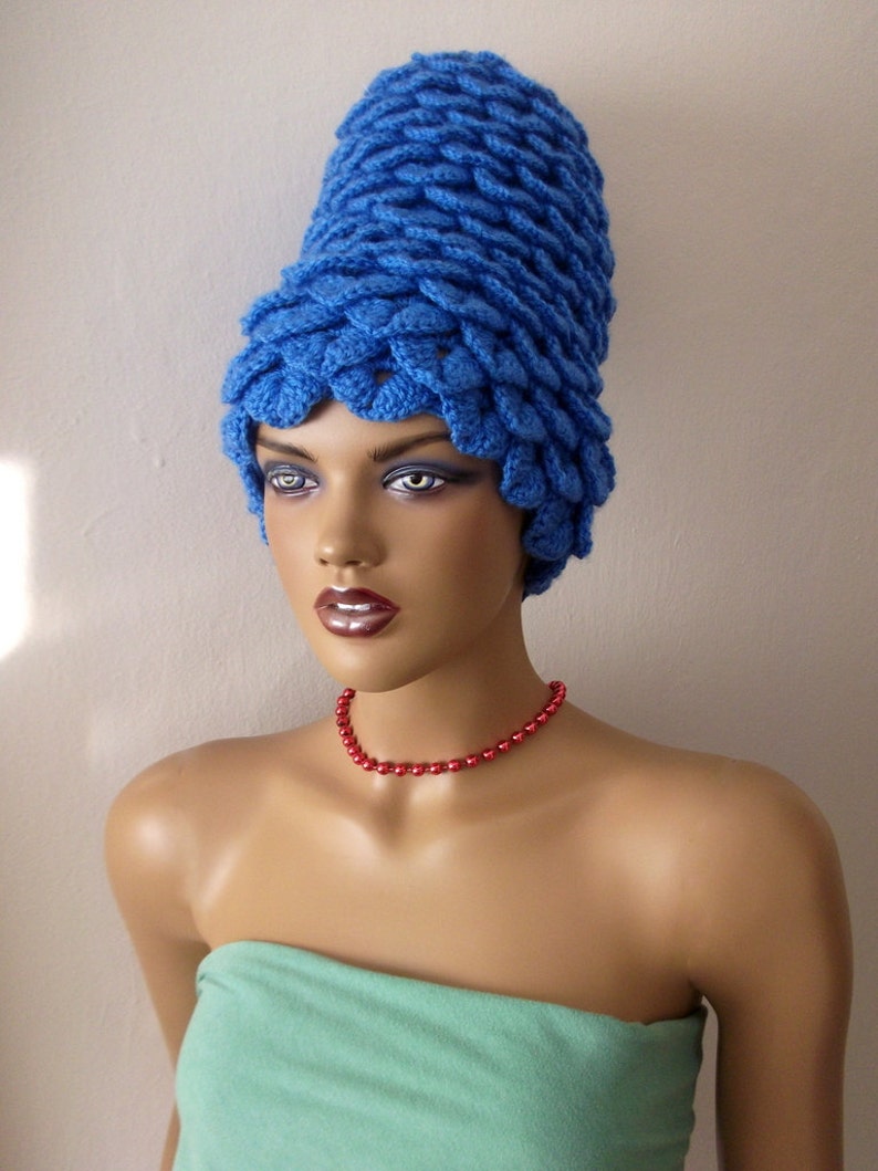 May include: A blue crocheted hat with a unique, pointed design. The hat is worn by a mannequin with a red beaded necklace.
