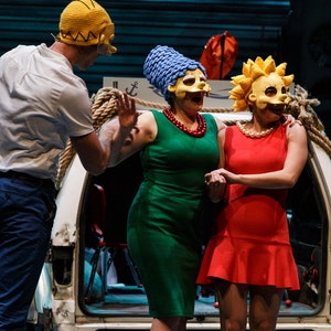 May include: Three people wearing yellow, blue and yellow masks, with the yellow masks having a cartoon face. The people are standing in front of a white van with a rope tied around it. The person in the blue mask is wearing a green dress and a red necklace. The person in the yellow mask is wearing a red dress. The person in the yellow mask is wearing a white shirt and blue trousers.