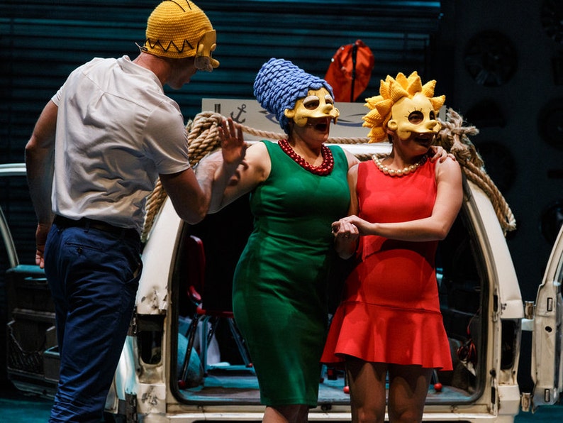 May include: Three people wearing yellow, blue and yellow masks, with the yellow masks having a cartoon face. The people are standing in front of a white van with a rope tied around it. The person in the blue mask is wearing a green dress and a red necklace. The person in the yellow mask is wearing a red dress. The person in the yellow mask is wearing a white shirt and blue trousers.