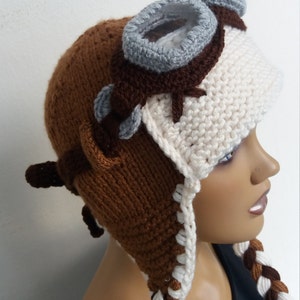 May include: Brown and white knitted hat with a gray crocheted aviator goggles design and long brown and white braided ear flaps.