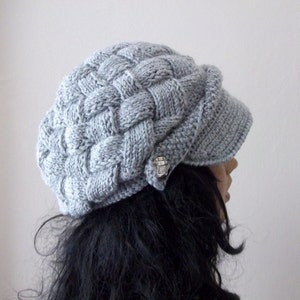 Könnte beinhalten: Eine graue Strickmütze mit einem geflochtenen Muster und einem kleinen silbernen Knopf an der Seite.