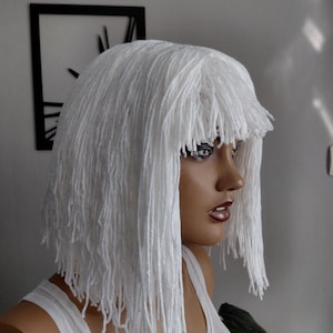 Puede incluir: Peluca blanca con un estilo corto y con flecos. La peluca está hecha de hebras largas y texturizadas, creando un efecto único y en capas. Se muestra en una cabeza de maniquí, mostrando su forma y textura. El maniquí lleva una camiseta blanca.