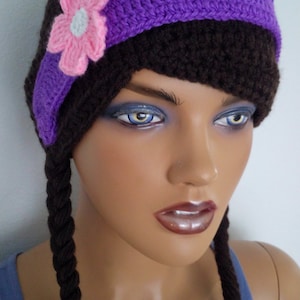 Puede incluir: Sombrero de punto marrón con una banda morada y una flor rosa. El sombrero tiene dos trenzas marrones largas unidas a los lados.