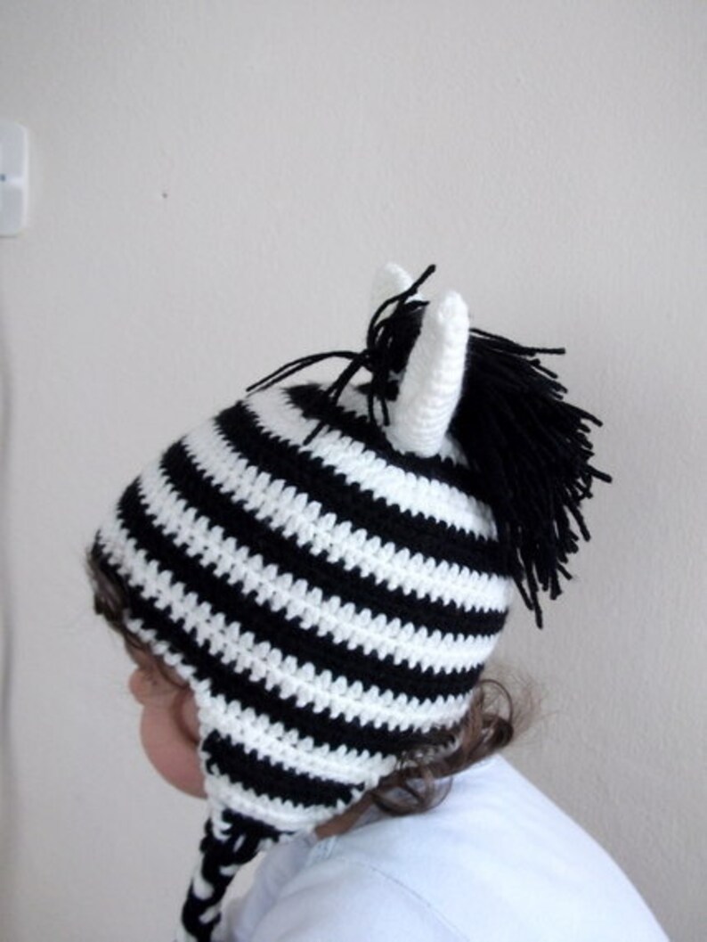 May include: A black and white striped crocheted zebra hat with a black pom-pom on top. The hat has long ear flaps.