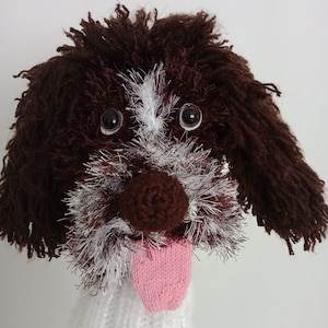 以下が含まれることがあります： 茶色と白の毛糸で編まれた犬の人形。ピンクの舌が突き出ています。人形は茶色の目と茶色の鼻をしています。