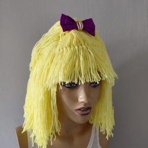 May include: A yellow yarn wig with a purple bow and gold-coloured ring detail. The wig is styled with a fringe and is displayed on a mannequin head. The mannequin is wearing a white vest top.