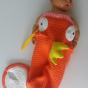 May include: A crocheted orange and white fish costume for a baby. The costume has a yellow tail fin and white eyes with black pupils.