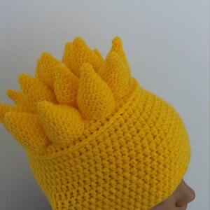 Puede incluir: Un gorro de crochet amarillo con un dise&ntilde;o de p&uacute;as. El gorro est&aacute; hecho con punto bajo y tiene una punta puntiaguda.