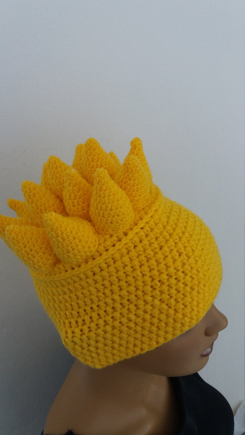 Puede incluir: Un gorro de crochet amarillo con un dise&ntilde;o de p&uacute;as. El gorro est&aacute; hecho con punto bajo y tiene una punta puntiaguda.