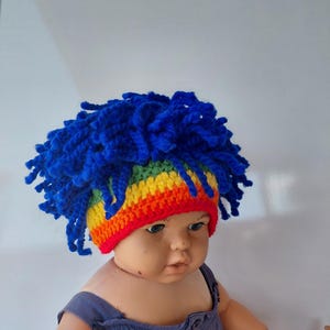 May include: A crochet hat featuring a vibrant rainbow-colored band in red, orange, yellow, and green, topped with a thick, voluminous mop of bright blue yarn strands that mimic a textured hairstyle, worn by a doll with a neutral expression.