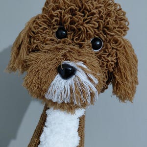 Puede incluir: Títere de perro tejido en marrón y blanco con ojos negros y nariz negra. El títere tiene un aspecto esponjoso y texturizado, con la cabeza y las orejas hechas de hilo marrón y un parche blanco en el pecho.