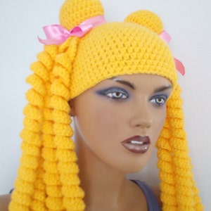 May include: A yellow crocheted hat with two long yellow crocheted pigtails and pink ribbons. The hat is worn on a mannequin head.