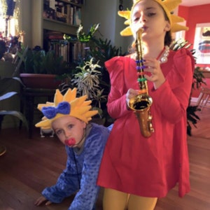 May include: Two children dressed in costumes. One child is wearing a yellow sun costume and playing a gold saxophone. The other child is wearing a blue costume with a yellow sun hat and a red pacifier.