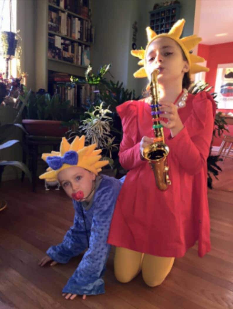 May include: Two children dressed in costumes. One child is wearing a yellow sun costume and playing a gold saxophone. The other child is wearing a blue costume with a yellow sun hat and a red pacifier.