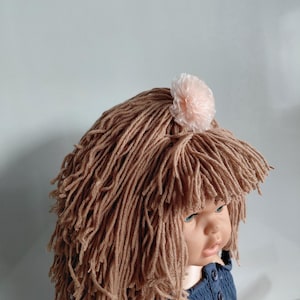 May include: A doll with a handmade brown yarn wig, featuring a light pink flower. The wig has long, shaggy strands. The doll is wearing a blue dress. The overall aesthetic is whimsical and playful.