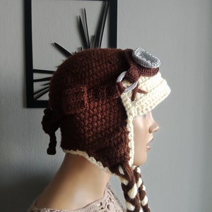 May include: Brown and cream crocheted aviator hat with a brown leather strap and a silver-colored goggle. The hat has long braids that are brown and cream.