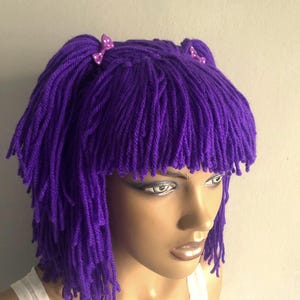 May include: A vibrant purple yarn wig featuring a fringe and two small pigtails, each adorned with a small bow with white polka dots. The wig is displayed on a mannequin head, highlighting the texture and colour.