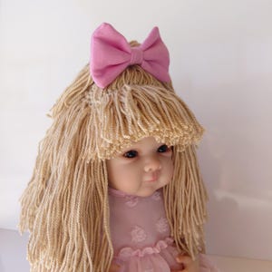 May include: A doll with light skin and dark eyes, featuring long, straight blonde yarn hair styled with bangs and a solid pink fabric bow on top. The doll wears a light pink dress with a textured, dotted pattern and ruffled layers on the bodice.
