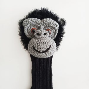 May include: A crocheted golf club head cover shaped like a monkey with black fur, gray face, and orange eyes.