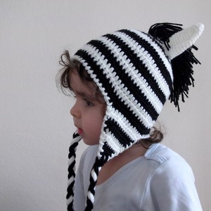 May include: A child wearing a black and white striped zebra hat with ear flaps and a black pom-pom on top.