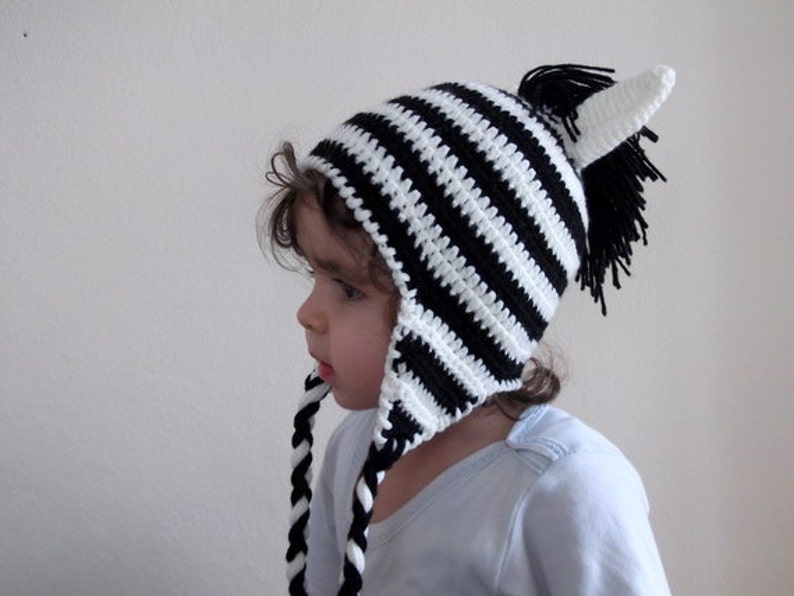 May include: A child wearing a black and white striped zebra hat with ear flaps and a black pom-pom on top.