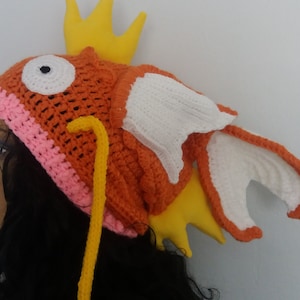 May include: A crocheted orange and pink hat shaped like a fish with a yellow fin and a white fin. The hat has a yellow crocheted antenna.