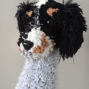 May include: A crocheted stuffed dog toy with black, white, and brown fur. The dog has a black nose and brown eyes.