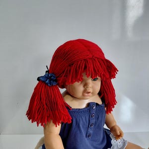 May include: A doll with red yarn pigtails and a navy blue dress. The doll's hair is styled in two ponytails, each secured with a blue ribbon. The dress has a button-up front and a white lace trim.