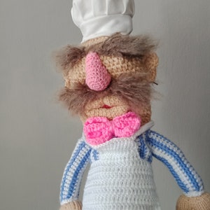 May include: A handmade crochet doll resembling a chef, with a white chef's hat, a pink nose, and a pink bow tie. The doll wears a white apron and a blue and white striped shirt. The doll has brown yarn hair and a beige body.