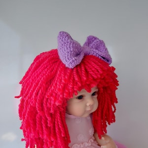 May include: A doll with bright red yarn hair styled in a layered cut, featuring a prominent purple crocheted bow attached to the top. The doll wears a light pink dress with a textured pattern and has a fair complexion with visible facial features.