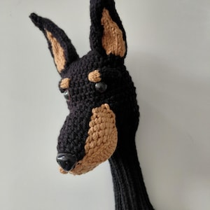 Könnte beinhalten: Eine schwarze und hellbraune Golfschläger-Kopfbedeckung aus Häkelarbeit in Form eines Dobermann Pinschers. Der Hund hat hellbraune Ohren und Schnauze, mit schwarzen Augen und einer schwarzen Nase. Die Hülle ist für einen Golfschlägerkopf konzipiert.
