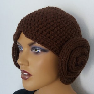 May include: Brown crocheted hat with two brown crocheted buns on the sides. The hat is worn on a mannequin head.