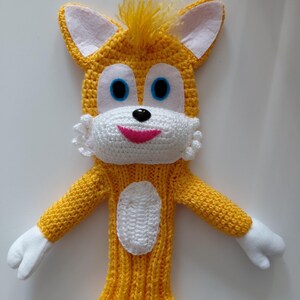 May include: A handmade yellow and white knitted character with blue eyes, a pink mouth, and white felt ears. The character has white felt hands and a white oval patch on the front. The top of the head has yellow tufts of yarn.