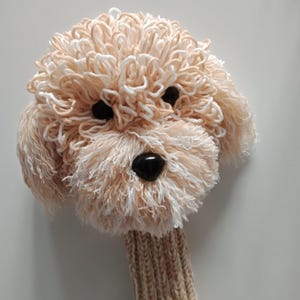 Puede incluir: Una funda para la cabeza del palo de golf con forma de perro, beige y blanca. La funda está hecha de hilo y tiene ojos negros y una nariz negra. El cuello de la funda es de punto beige.