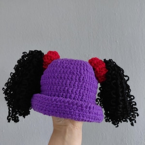 May include: A purple crocheted hat with black curly hair extensions and red accents. The hat is handmade and features a unique design, suitable for costume or everyday wear. A fun and whimsical accessory.