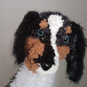 May include: A handmade plush dog with black, brown, and white fur. The dog has blue eyes and a black nose. The plush toy is made of soft, textured materials, and the dog's ears are long and floppy.
