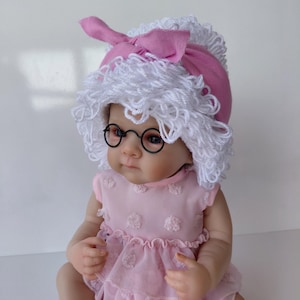 Peut inclure: Une poupée portant une robe rose avec un motif à pois texturé et une jupe à volants. La poupée porte un bonnet en laine blanche avec un nœud en tissu rose sur le dessus et de petites lunettes rondes à monture noire. Elle porte aussi des chaussettes roses.