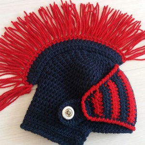 May include: A navy blue crocheted hat with a red fringe, resembling a Roman gladiator helmet. The hat features a side piece with red and navy stripes and a decorative button. This handmade item is suitable for costume or play.