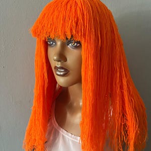 May include: A mannequin head with a bright orange wig. The wig has a blunt fringe and long, flowing strands. The mannequin is wearing a white top. The background is a plain grey.