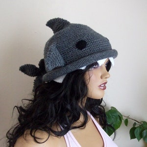 May include: Gray knitted shark hat with white teeth and black eyes. The hat is worn by a mannequin with long black hair.