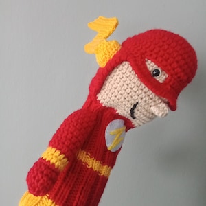 May include: A handmade, crocheted Flash golf club head cover. The cover is primarily red with yellow accents, including a lightning bolt emblem and a yellow lightning bolt on the head. The face is tan with black eyes and a smile.